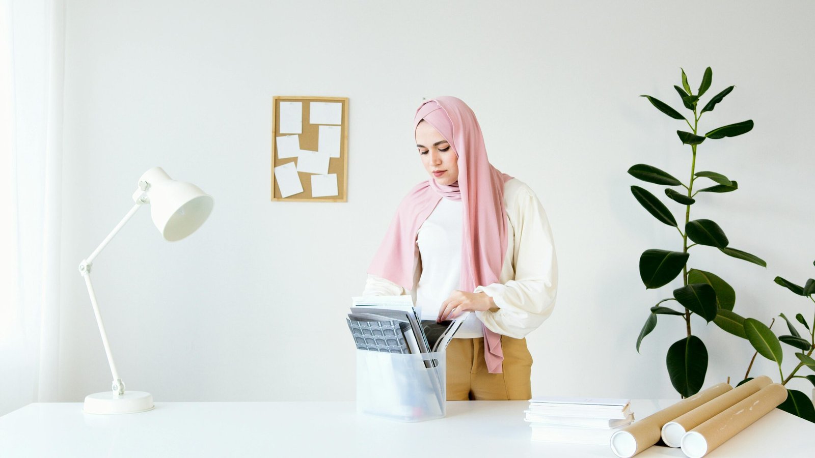 A woman wearing a hijab stands at a desk, organizing documents in a modern, minimalist office setting.