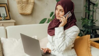 A Muslim woman in a hijab working remotely, balancing a phone call and laptop in a home office setting.