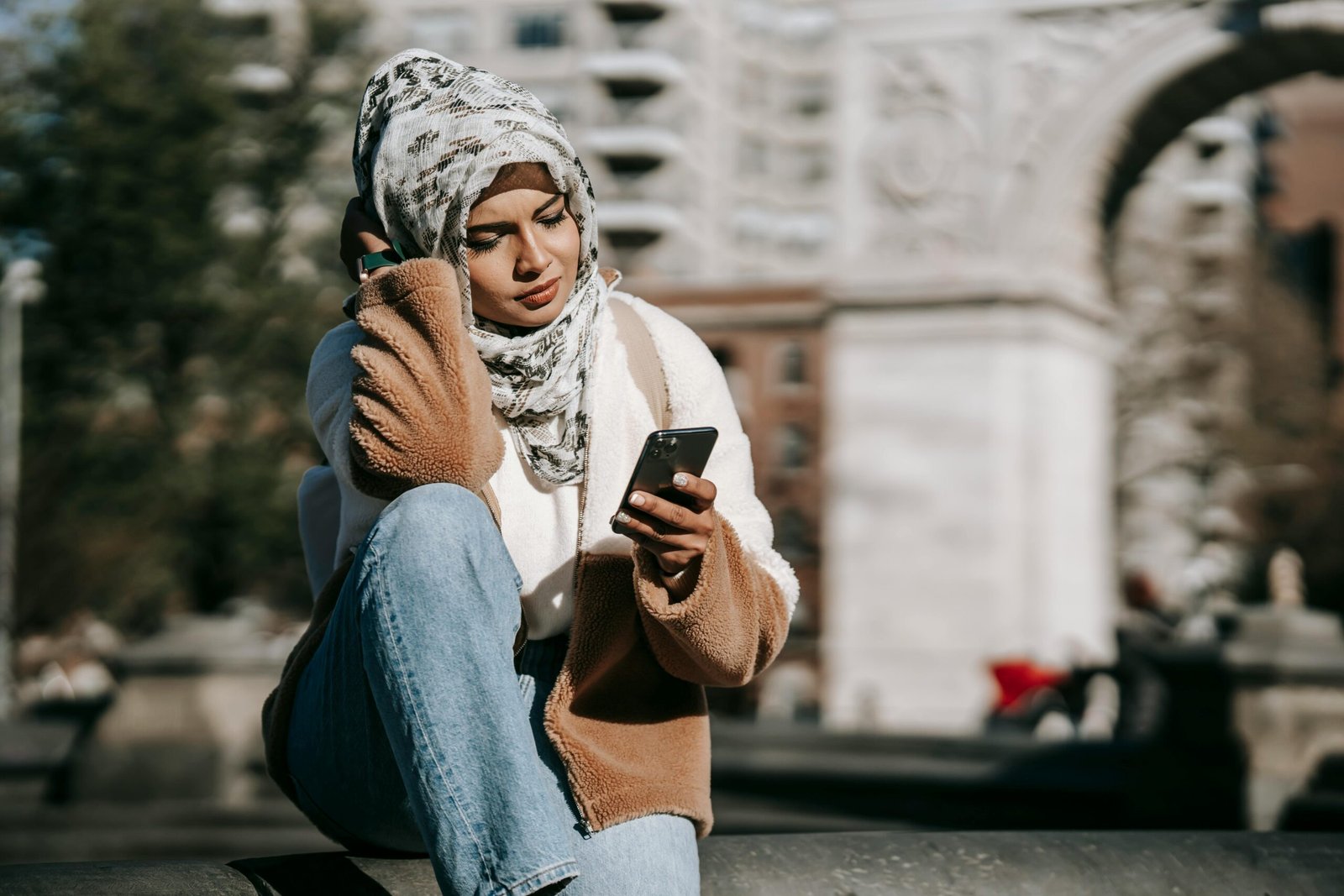 Stylish young Arab female in casual warm outfit and traditional hijab resting on city square and messaging on mobile phone on sunny day